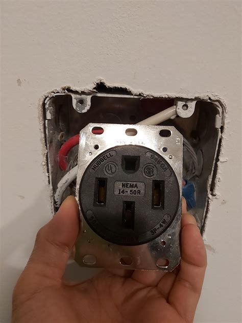 Hand holding a black NEMA 14-50R outlet, mounted in a wall box with red, white, and blue wires.