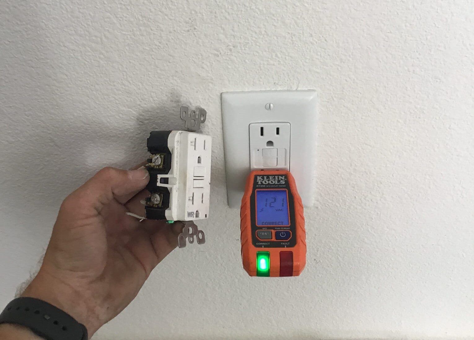Hand holding electrical outlet next to wall outlet with electrical tester displaying a reading.