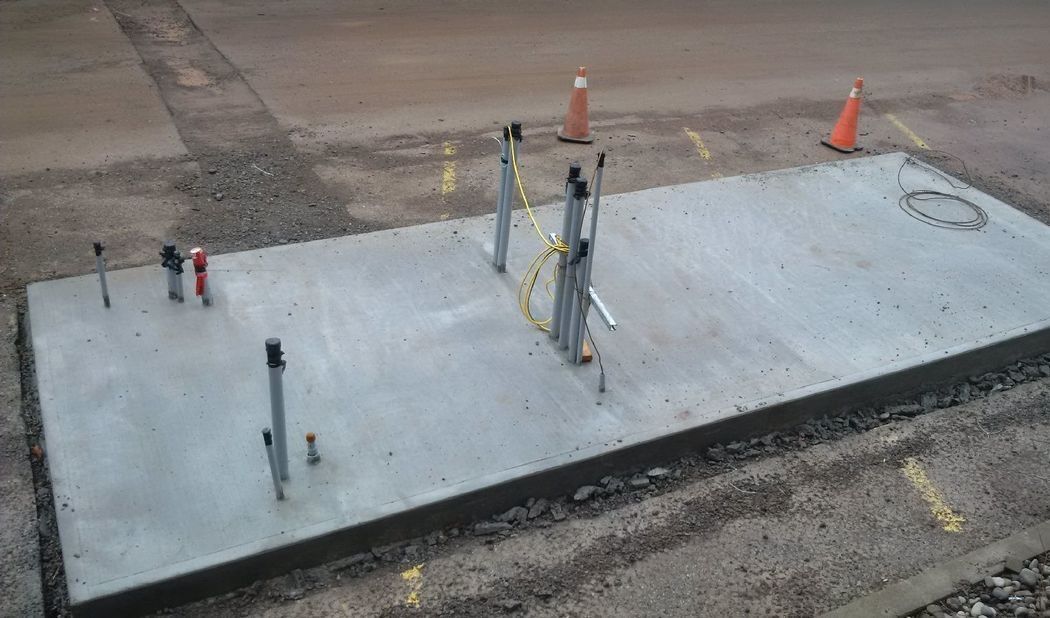 Concrete pad with metal poles and caution tape, likely for utilities. Orange cones and markings in road.