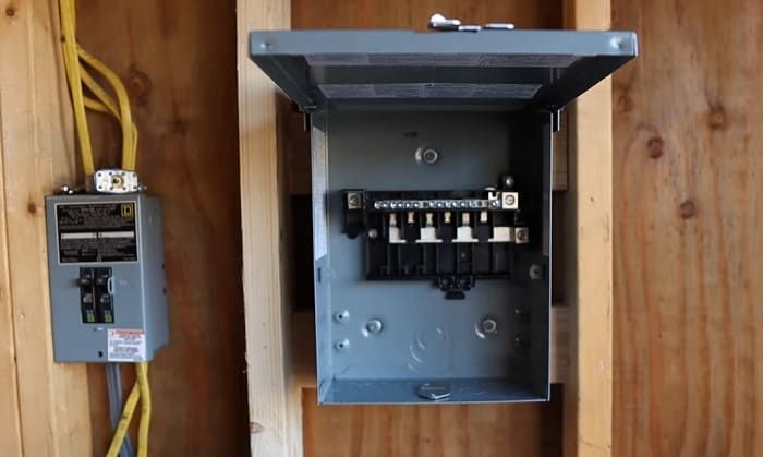 Electrical panel with the cover open, mounted on a wooden wall next to a breaker box.