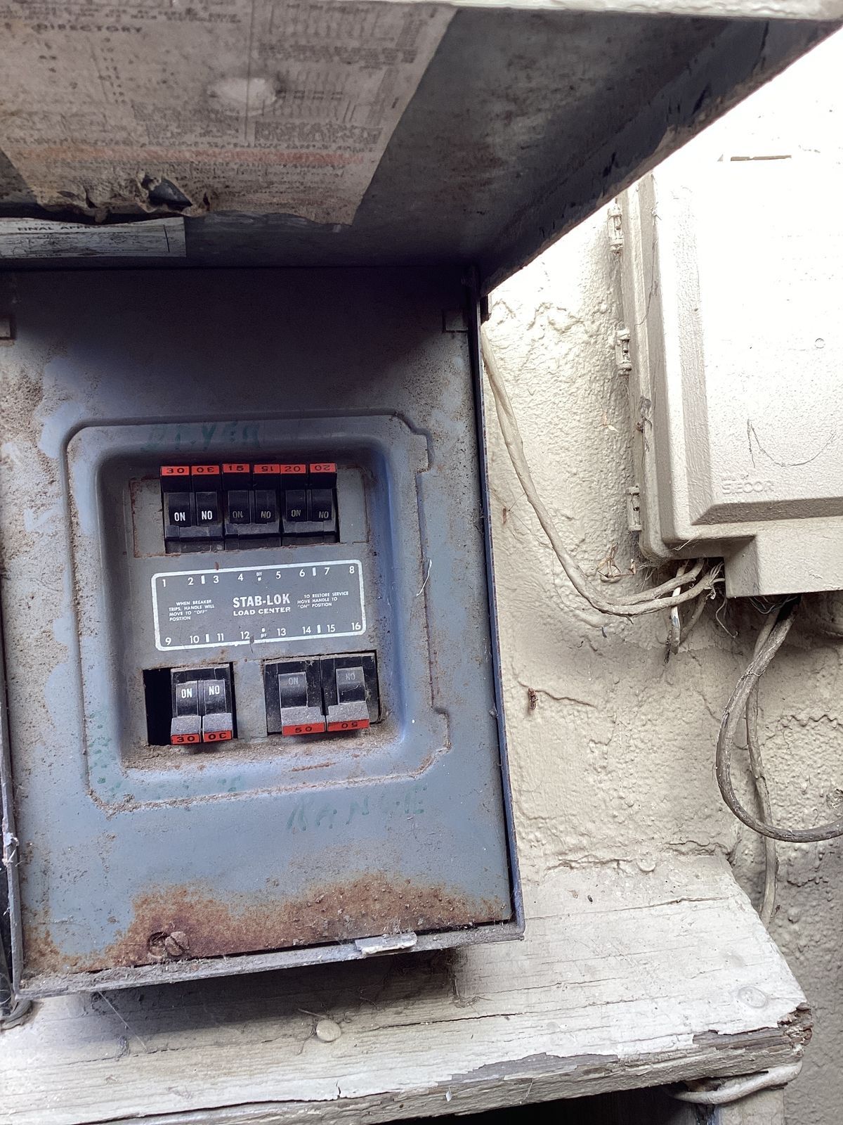 Old, gray electrical panel with rust, housing circuit breakers on a weathered wall.