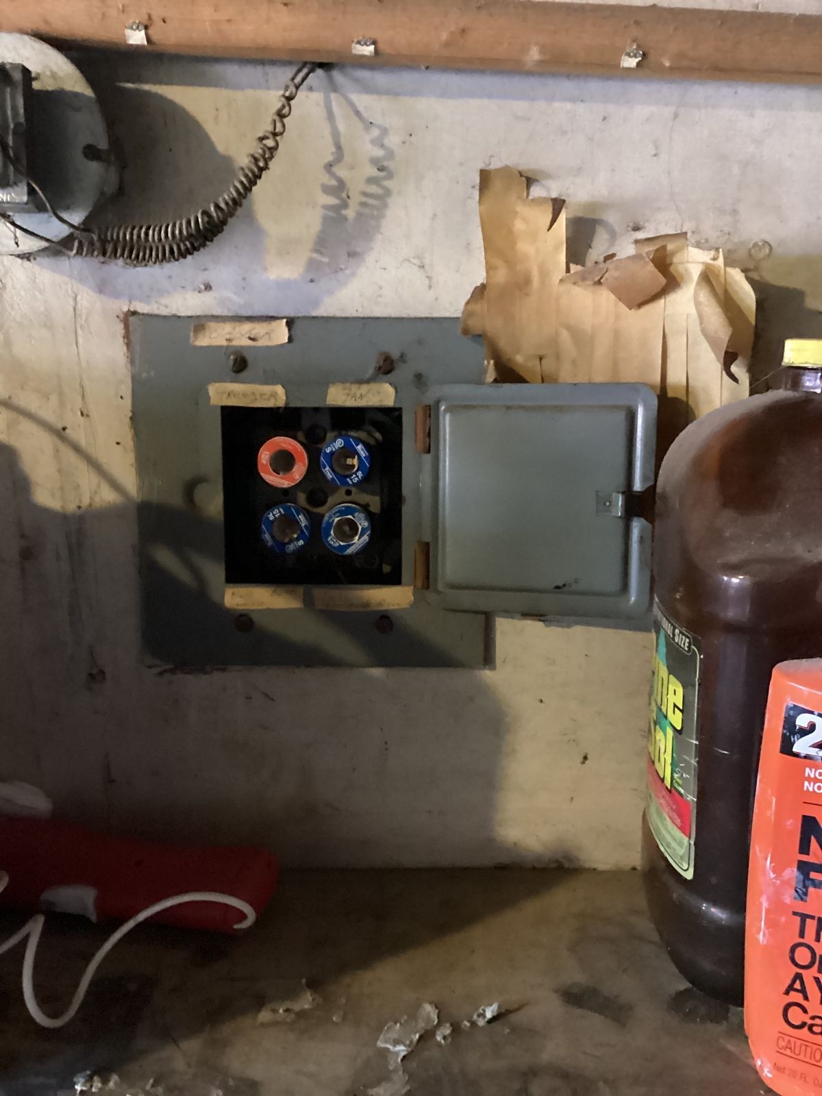 Open electrical fuse box on a concrete wall. Wires and debris are visible. A brown bottle is to the right.
