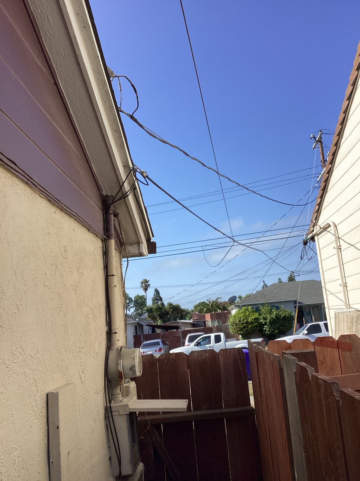 Overhead power lines cross a blue sky between two buildings. A wooden fence is in the foreground.