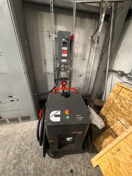 Cummins generator in a utility room, gray and black, connected to electrical panel with red and green indicator lights.