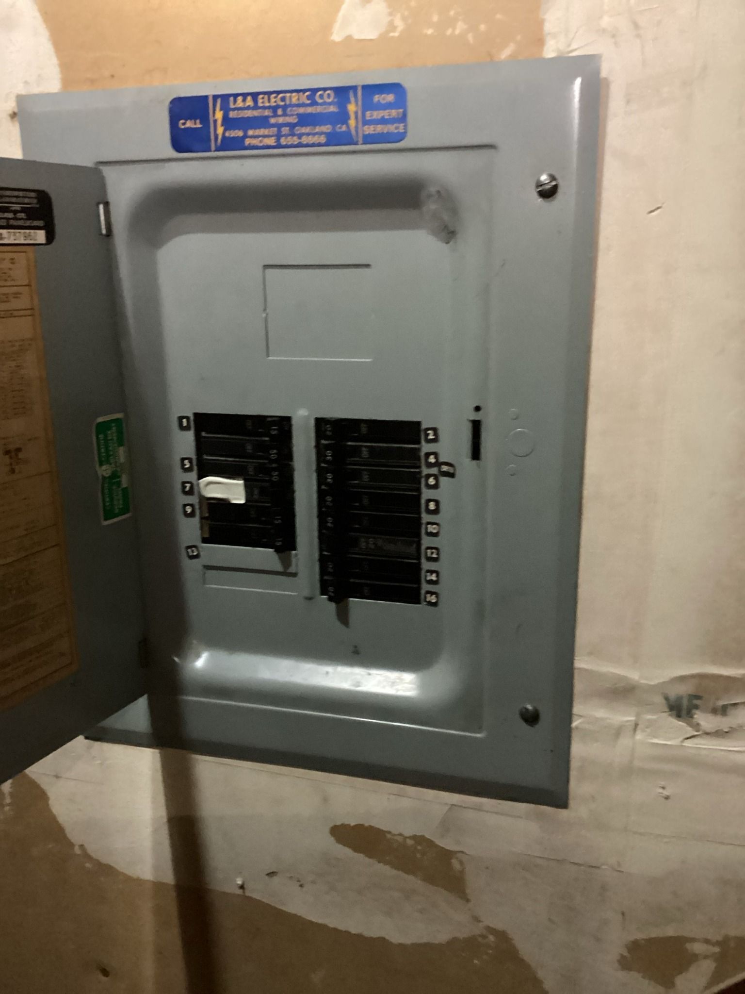 Opened gray electrical circuit breaker panel on a rough wall. Labels and breakers visible.