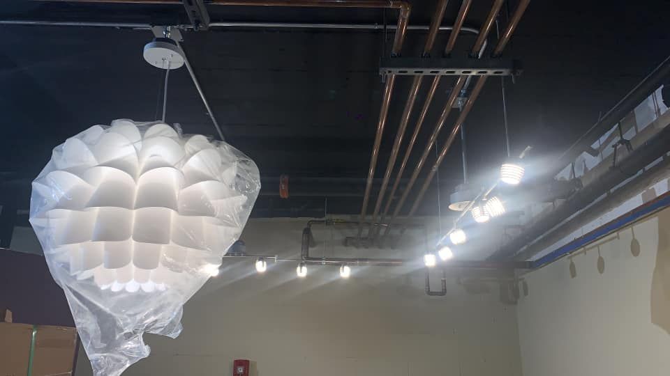 A decorative white paper lantern hangs, along with a string of lights, in a room with exposed pipes and a black ceiling.