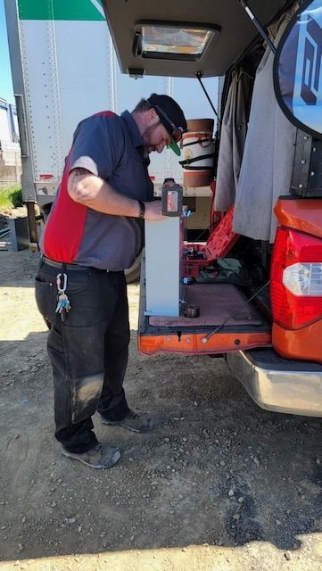 Mechanic working on equipment near a truck, wearing safety glasses and uniform, outside in daylight.
