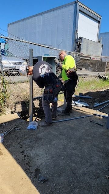 Two workers installing a metal fence post outdoors, sunny day. One in a safety vest.