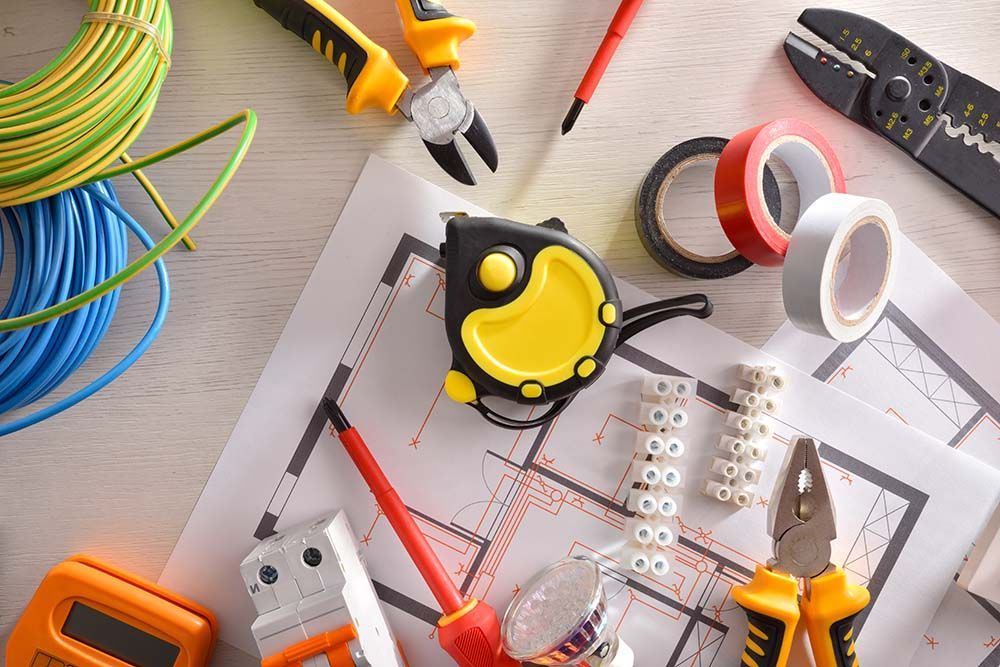 Electrical tools, wiring, and blueprints on a light wood surface. Includes tape, wire strippers, and a multimeter.