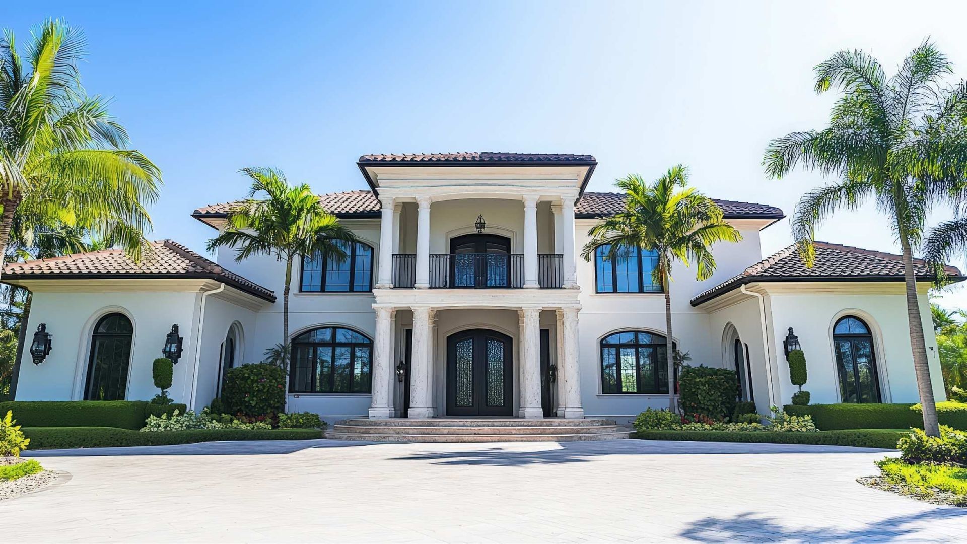 White mansion with columns, black windows, and palm trees under a bright blue sky.