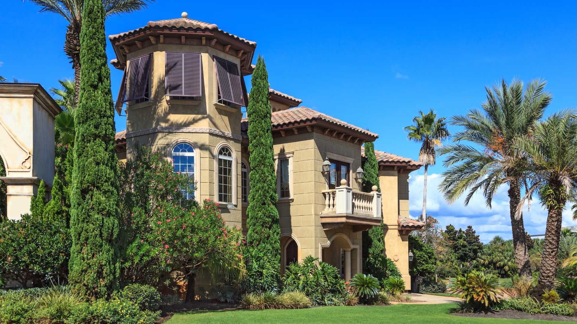Mediterranean-style house with a tower and balcony, set against a blue sky with palm trees.