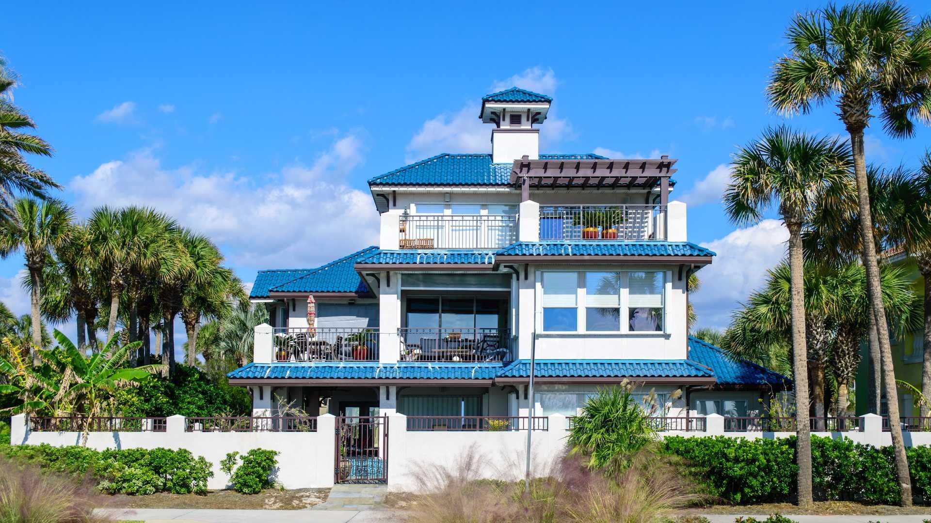 White multi-story house with blue tiled roof and balcony surrounded by palm trees.