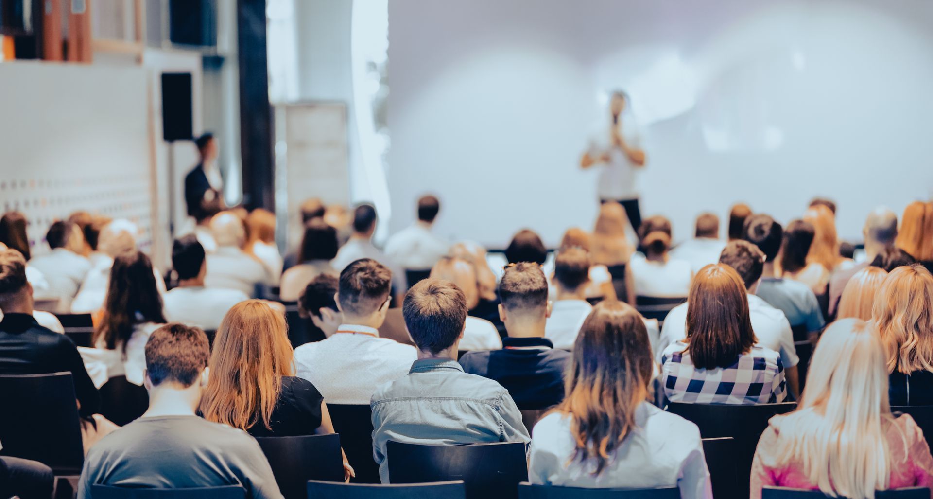 Público en una sala de conferencias escuchando a un orador en un podio frente a una pantalla de proyección.