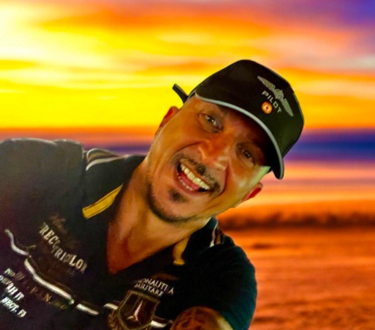 Un hombre que lleva un sombrero de piloto sonríe ampliamente frente a un atardecer de fondo.