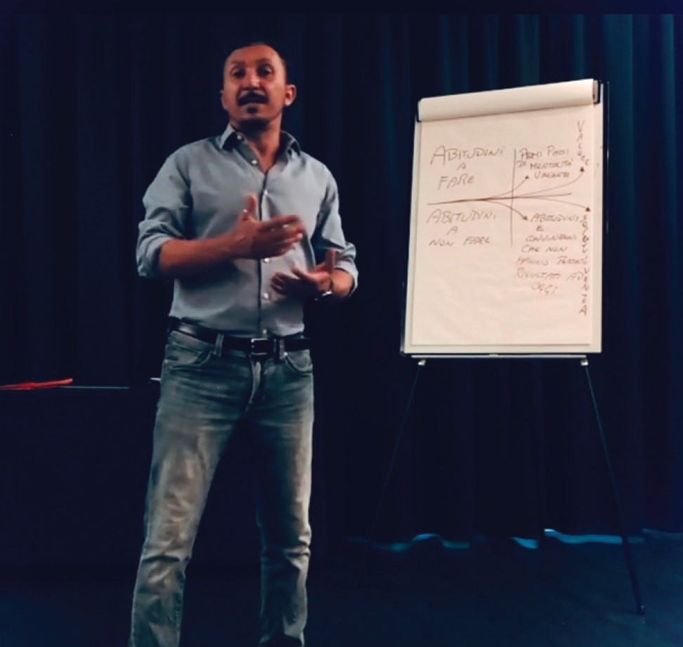 Uomo che gesticola, mentre presenta qualcosa vicino a una lavagna con appunti scritti a mano. Sfondo scuro.