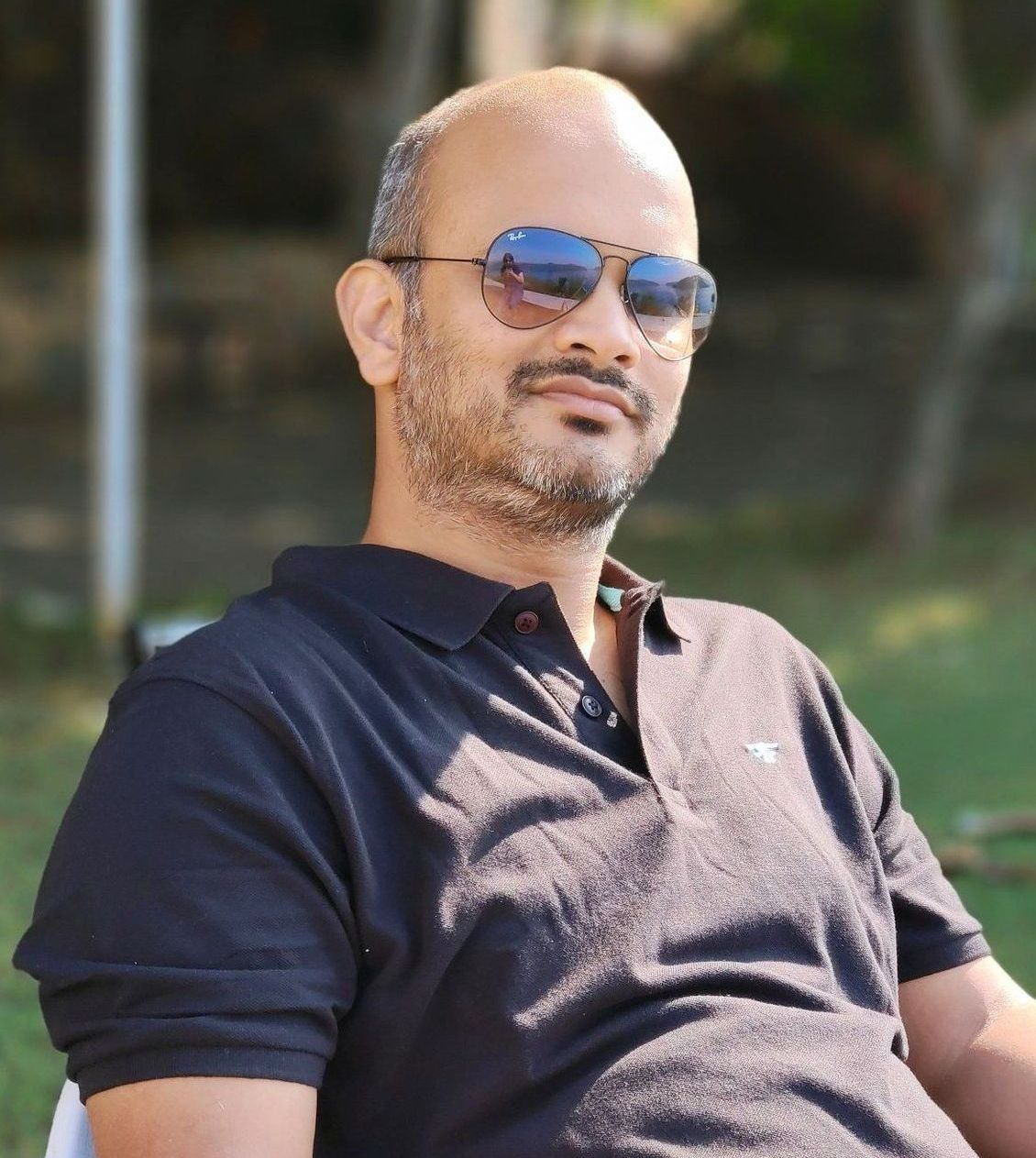 Bald man in sunglasses, wearing a black polo shirt, outdoors, with blurred greenery in the background.