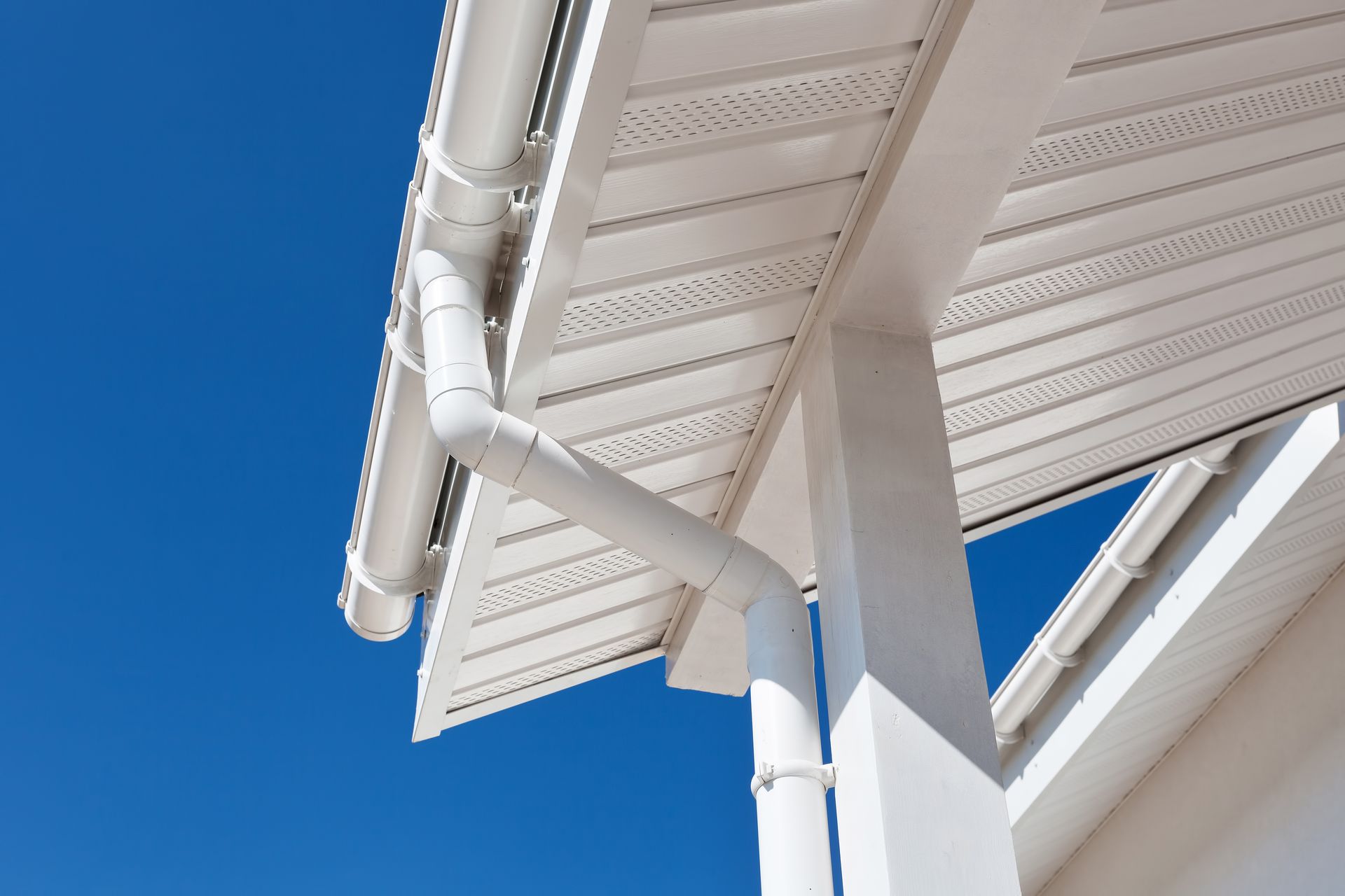 White house gutter system against a clear blue sky. The downspout curves down a support beam.