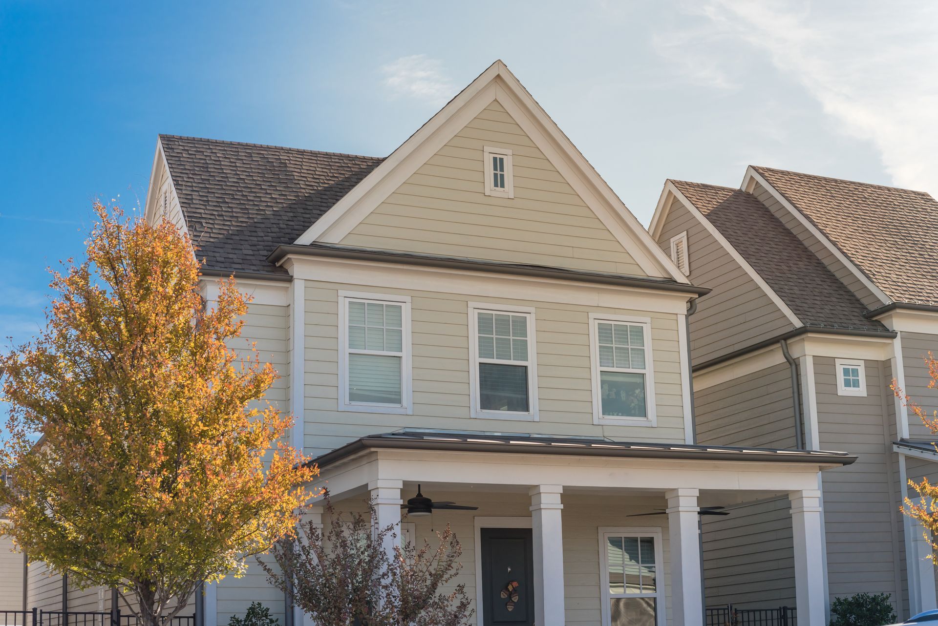 Two-story beige house with a porch and columns; fall tree in the foreground, blue sky.