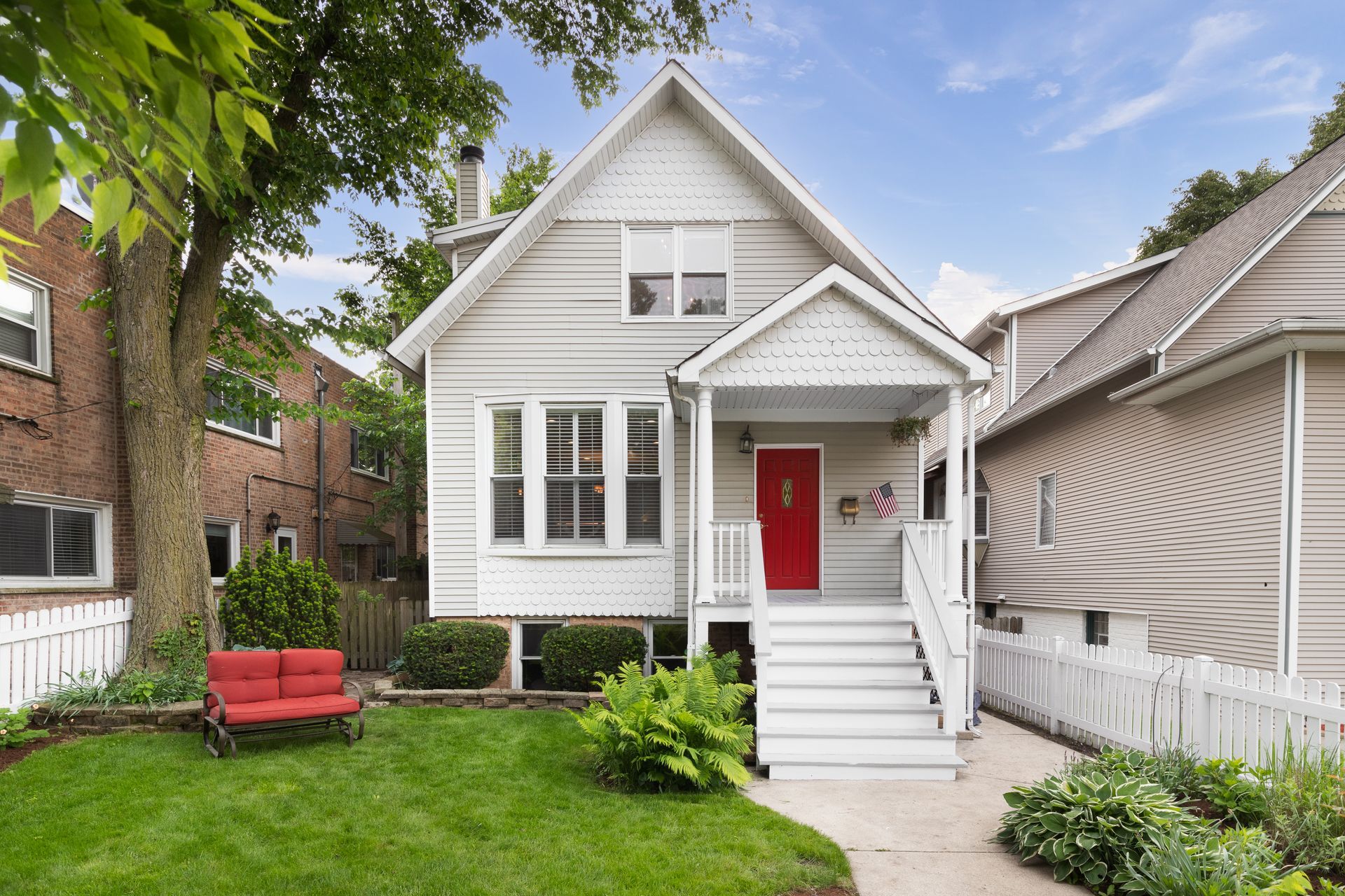 Gray clapboard house with a red front door, white porch, and picket fence, with a small lawn and tree in the front yard.