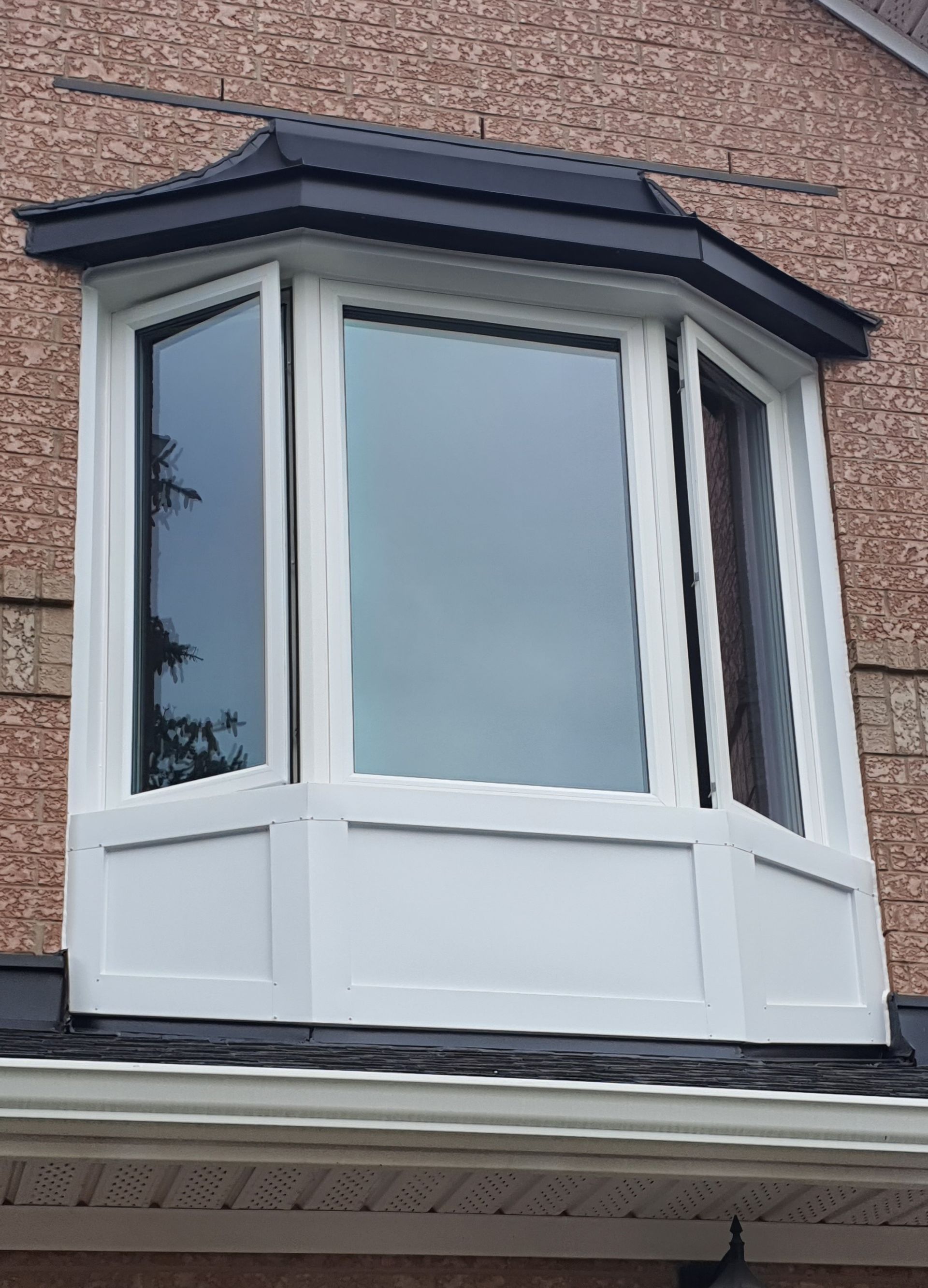 Bay window with white trim and a dark roof against a brick exterior.