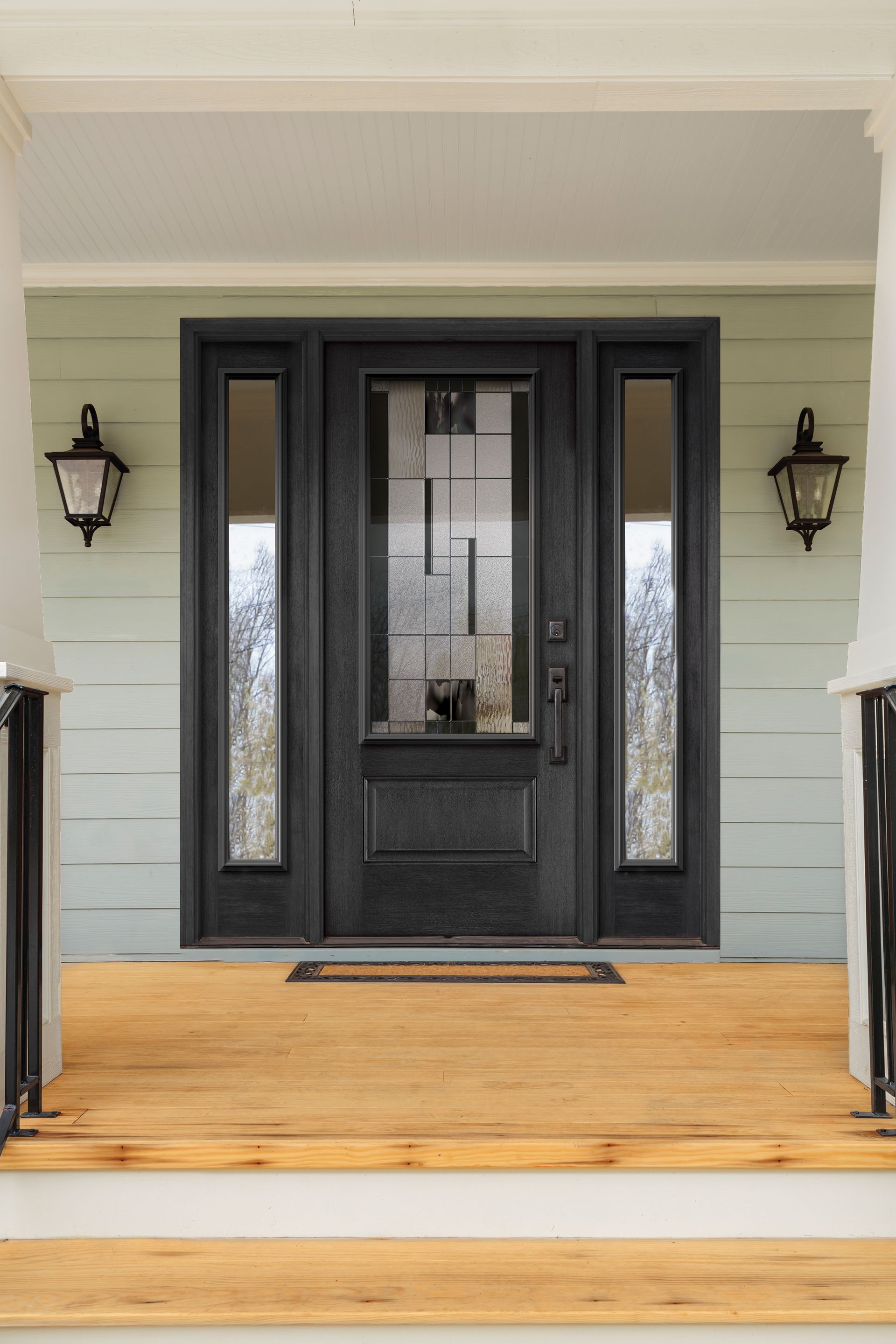 A black front door with sidelights on a wooden porch, flanked by wall-mounted lanterns. The door has a decorative glass panel.