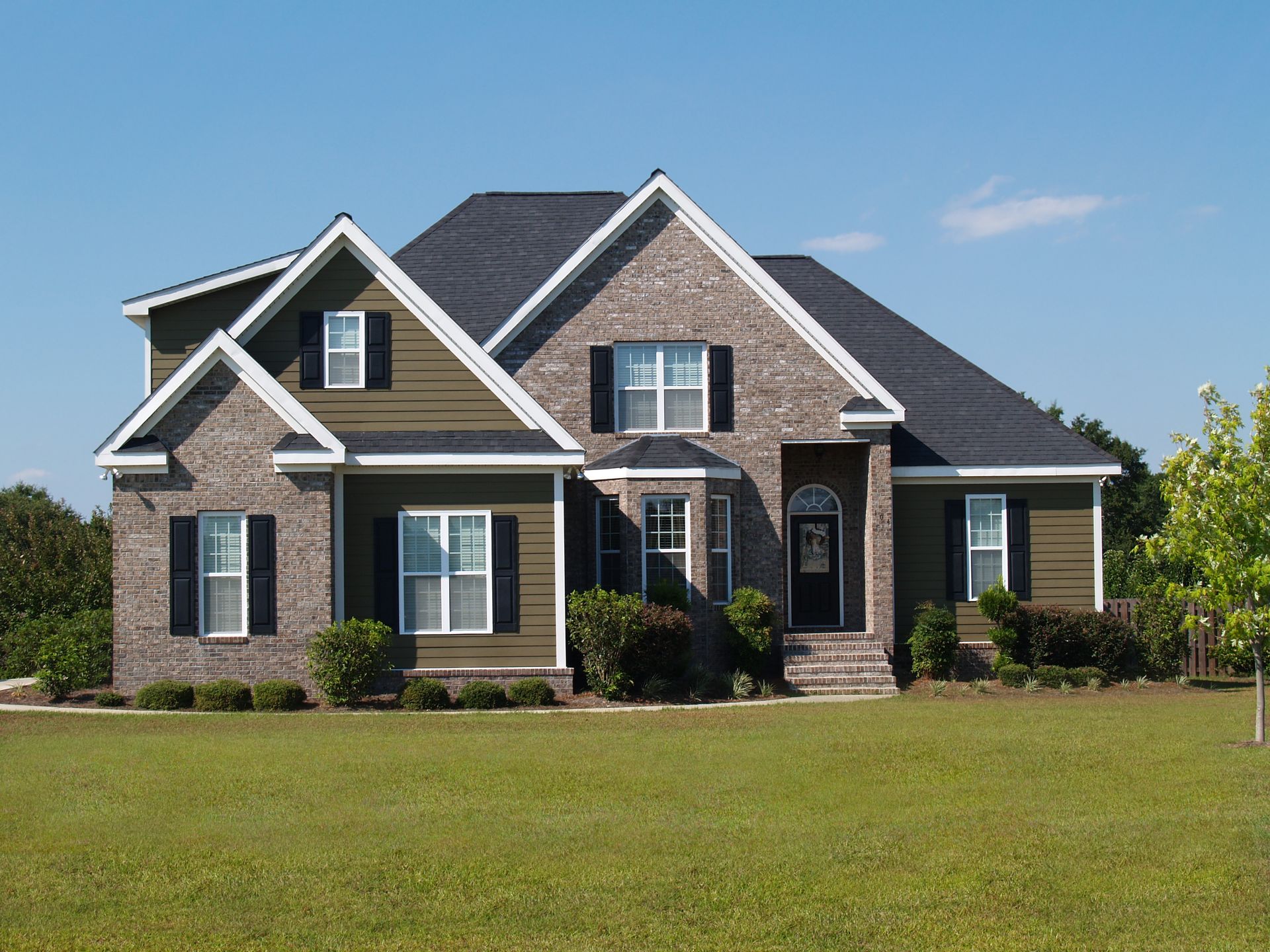 Two-story house with green siding, brick accents, black shutters, and a well-manicured lawn under a blue sky.