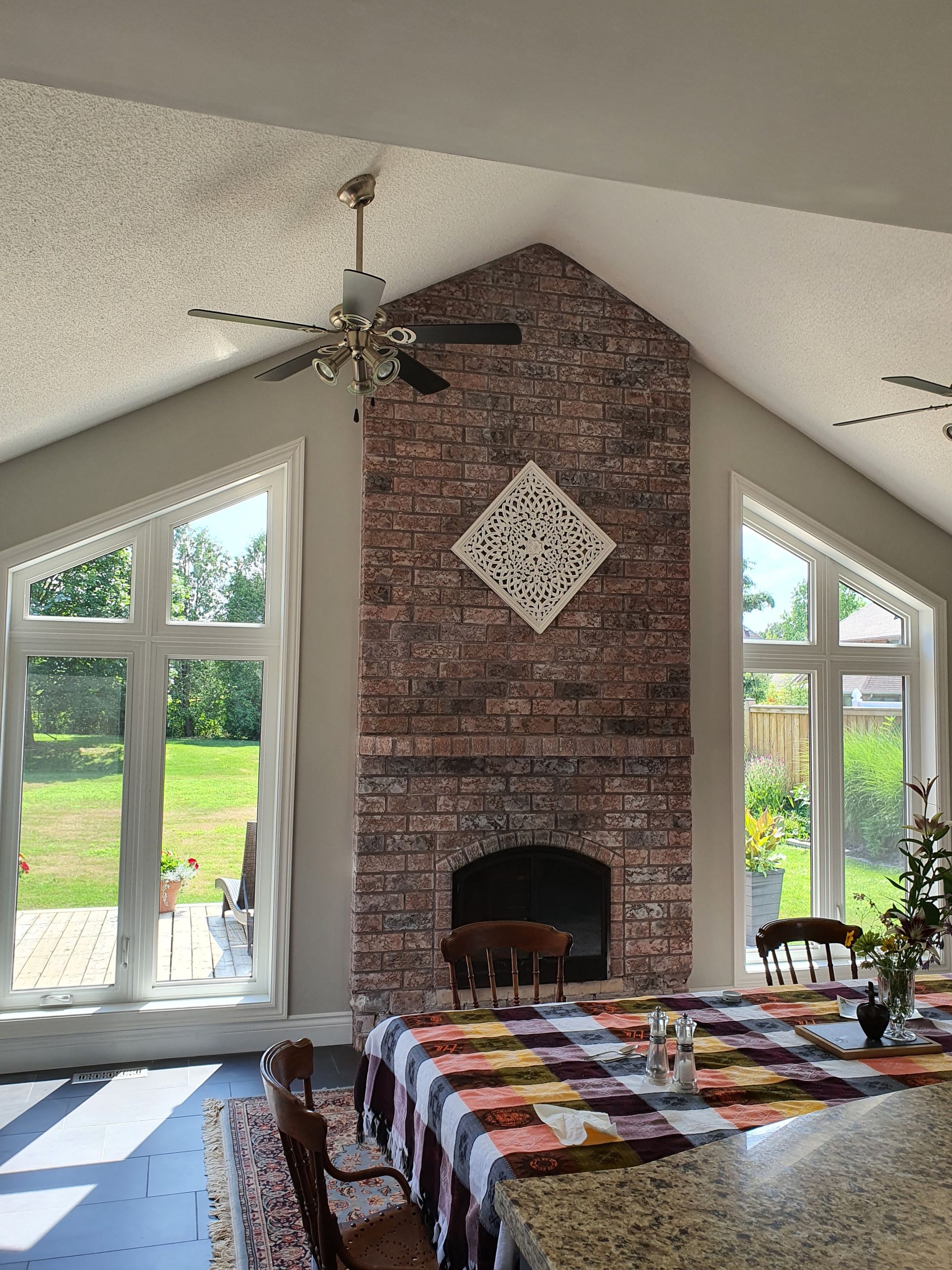 A brick fireplace with a white decorative piece sits between two large windows in a sunlit room. A table with a checkered tablecloth is in front.