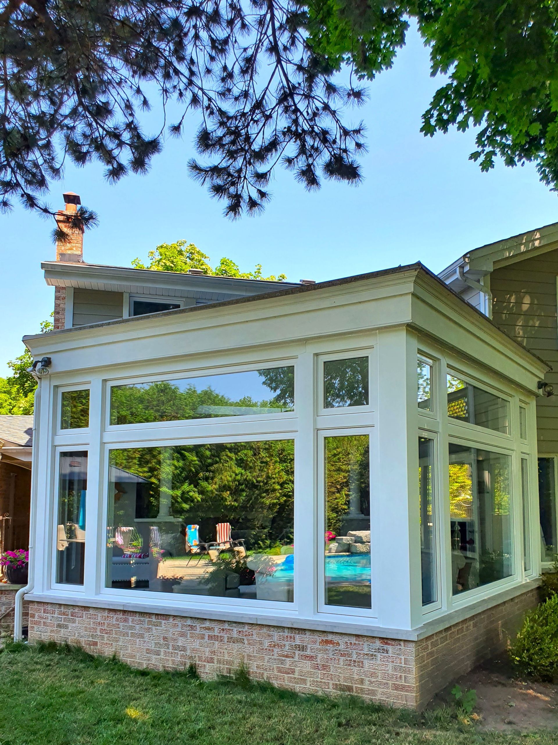 White sunroom with large windows attached to a two-story house, situated on a grassy lawn with a pool visible through the windows.