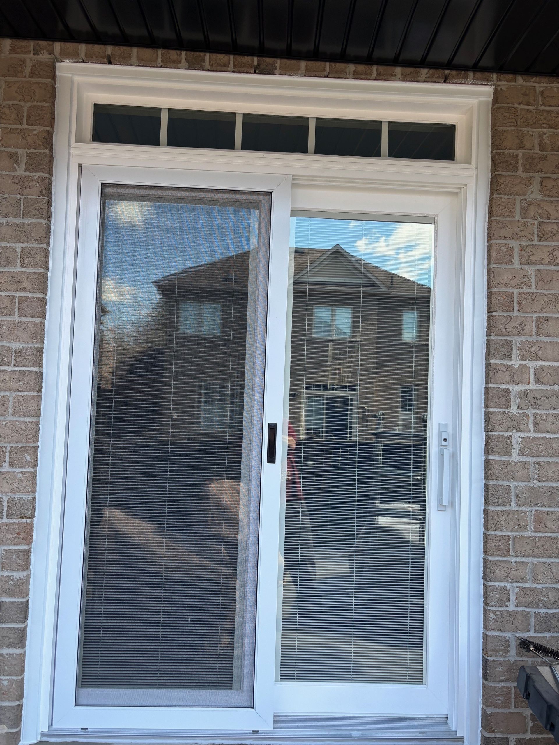 Sliding glass door with white frame and horizontal blinds reflecting a house. Brick exterior.