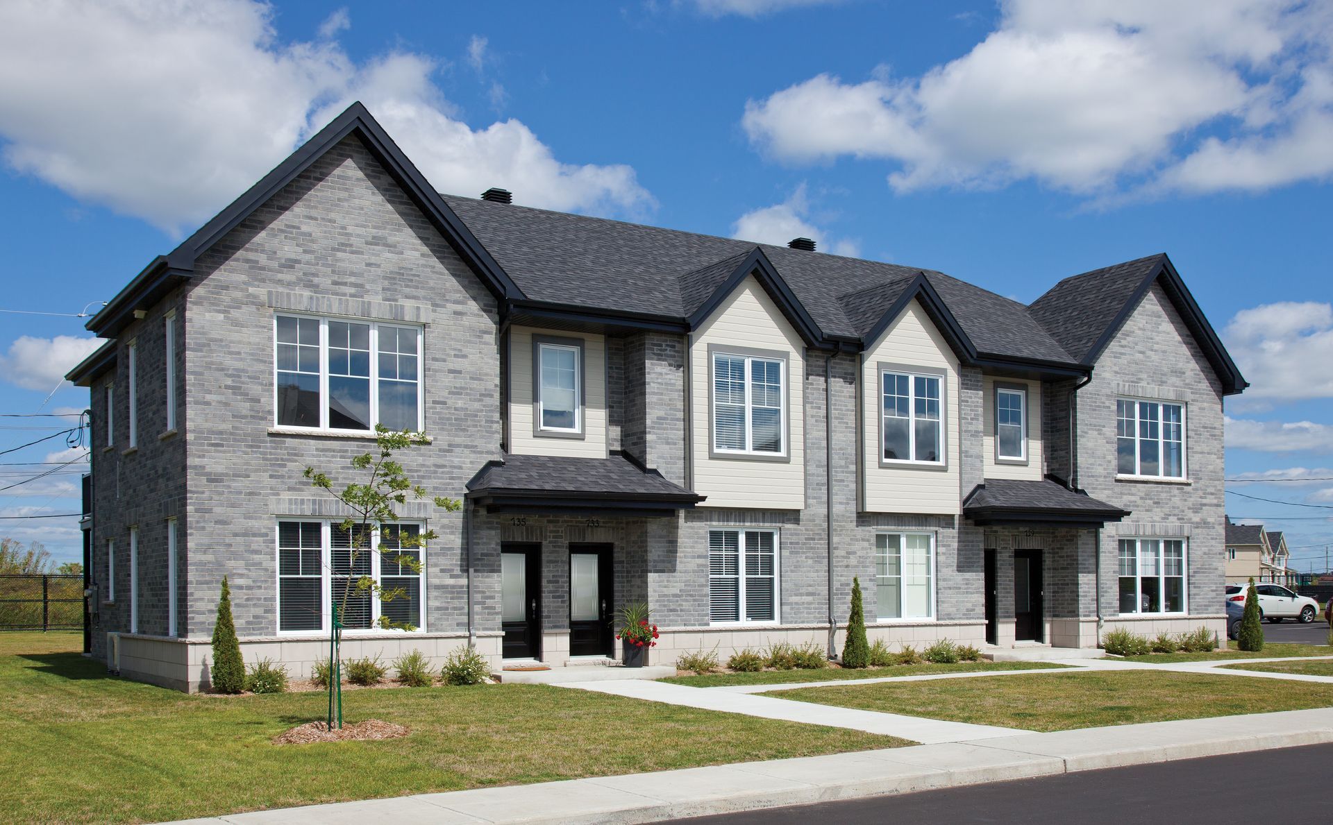 Gray brick townhouses with black roofs and white window frames against a blue sky. Small lawns in front.