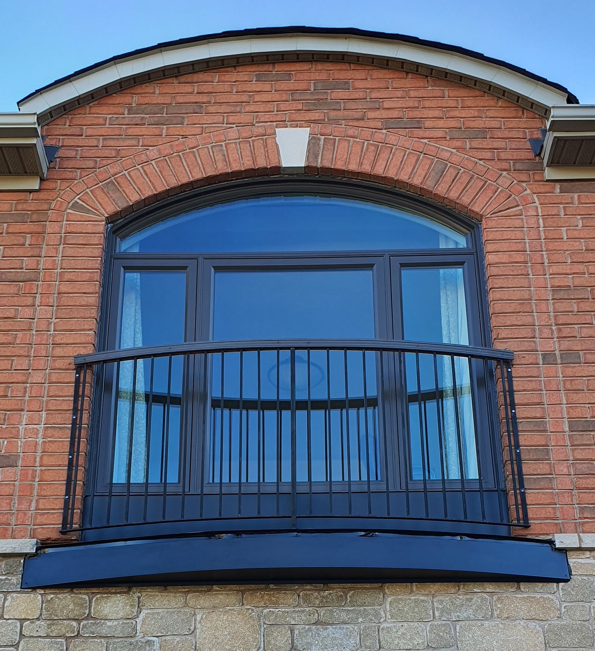 A dark-framed, arched window with a black metal balcony railing set into a red brick wall with a stone base.