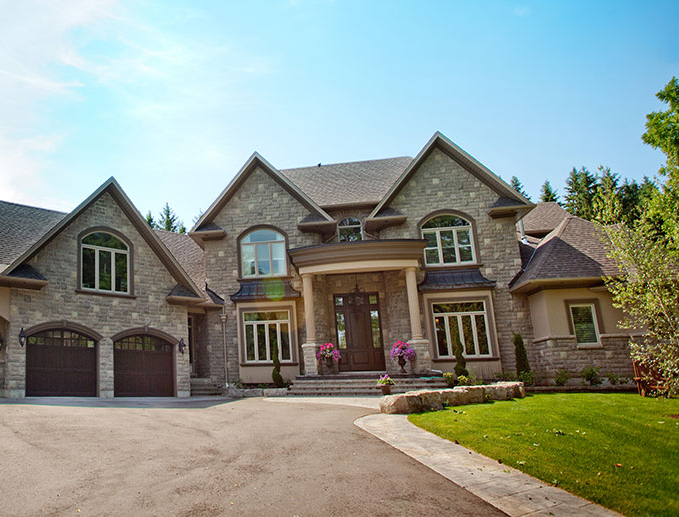 Large, multi-story stone luxury house with a paved driveway, two-car garage, and green lawn under a clear blue sky.