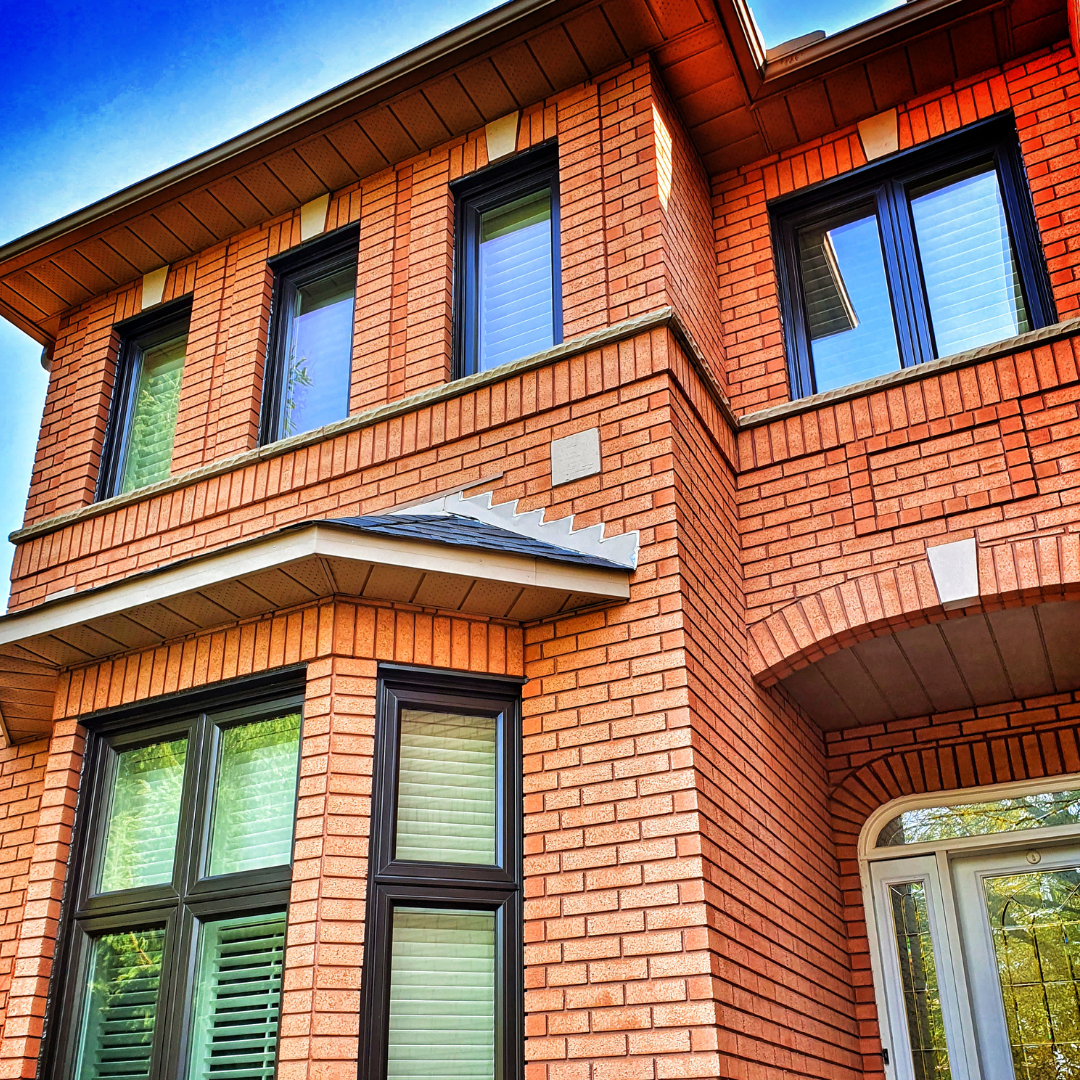 A two-story brick house facade with dark-framed windows and an arched entryway under a bright blue sky.