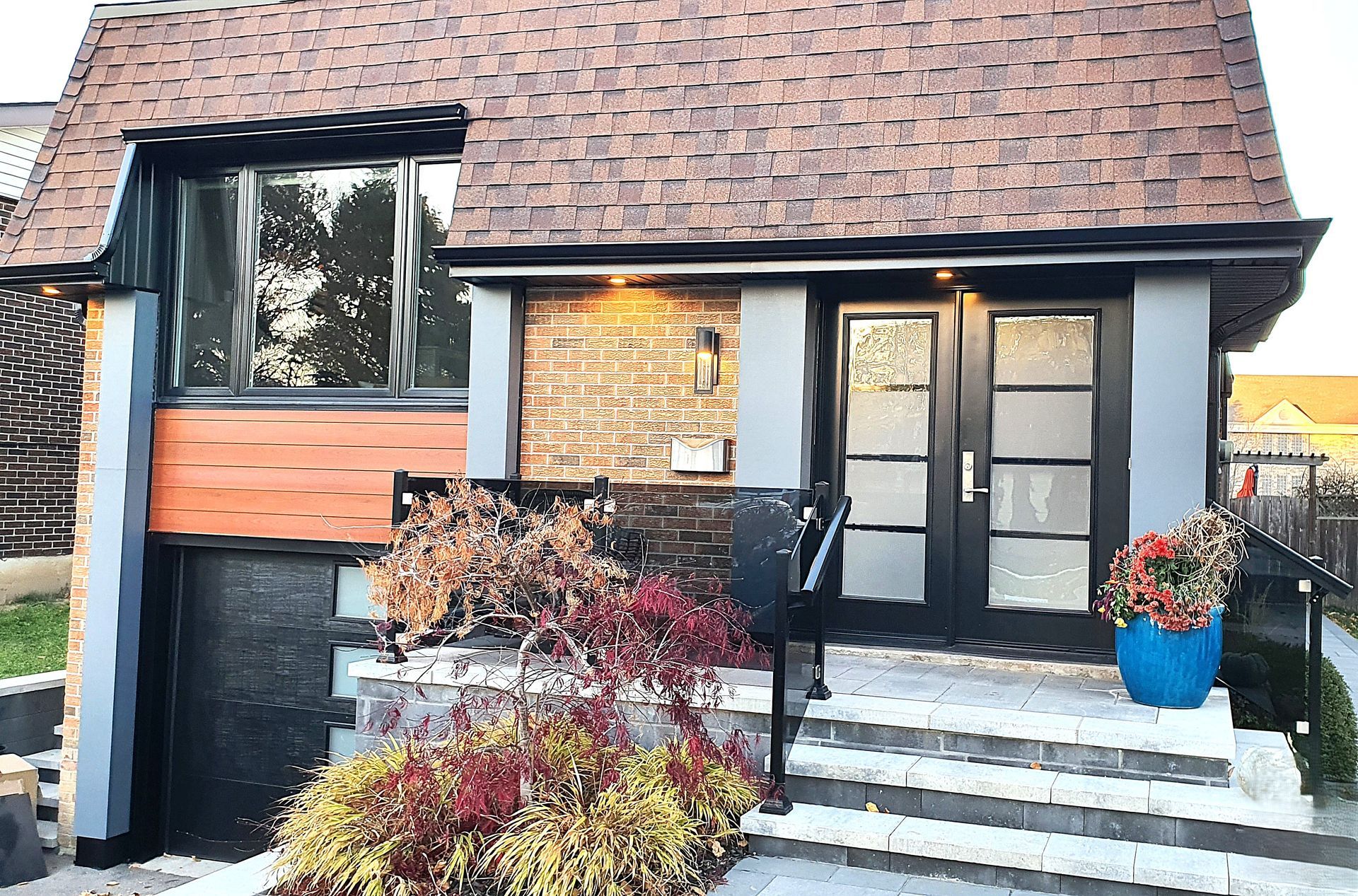 A front view of a house with a shingled roof, brick facade, black double doors, a grey staircase, and a potted plant.