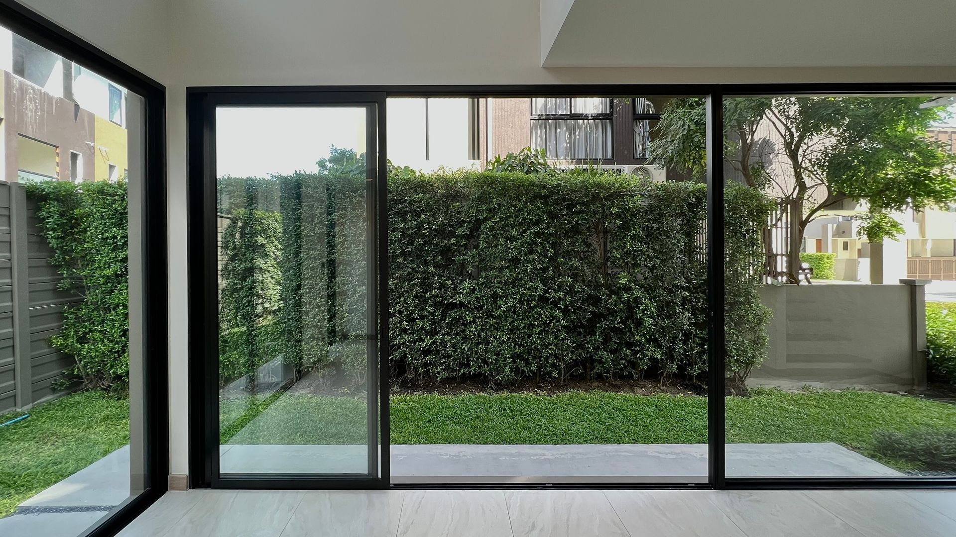Large floor-to-ceiling sliding glass doors look out onto a green backyard with tall hedges and a light-colored fence.