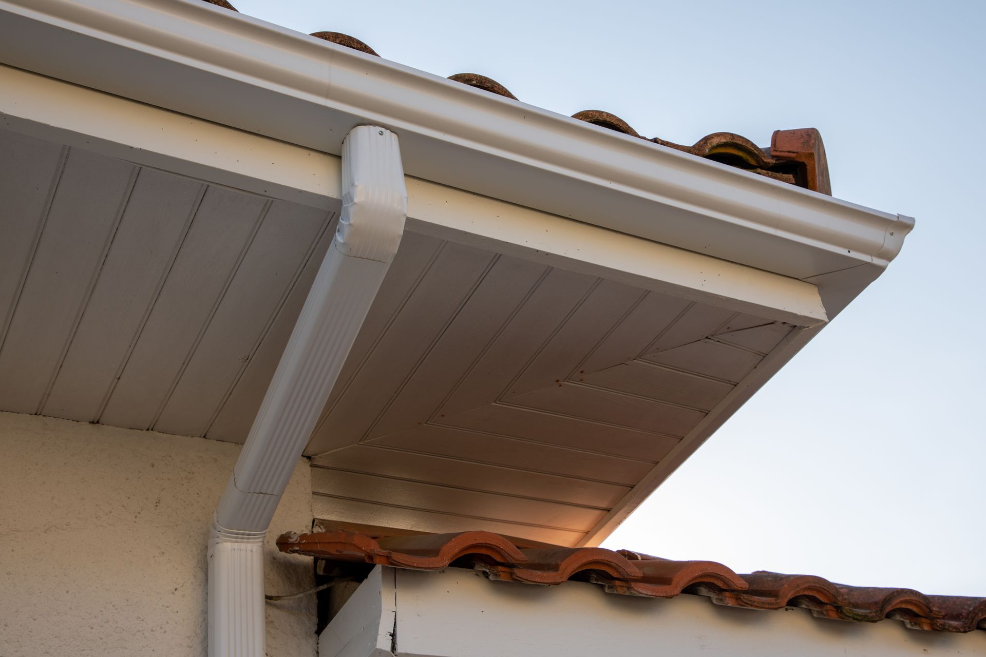 White rain gutter and soffit on a house with a terracotta tile roof, against a blue sky.