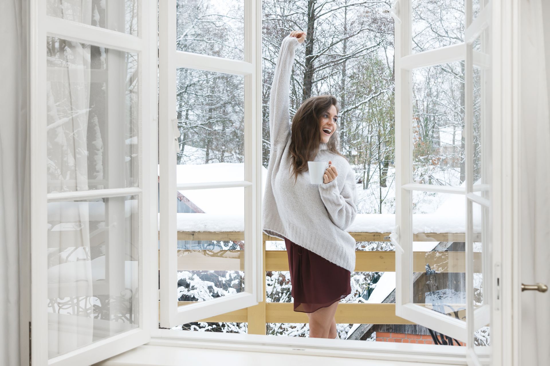 Woman in sweater on snowy balcony, arms raised, by open white windows.