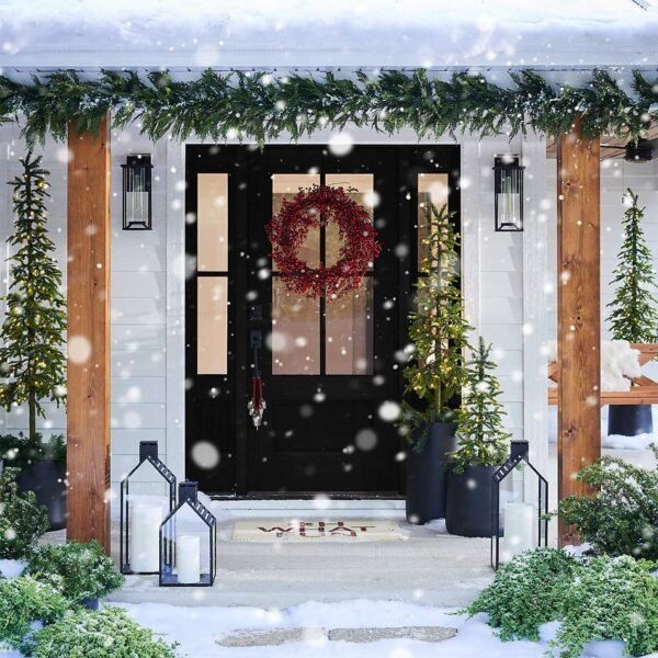 Snowy Christmas front porch, black door with wreath, greenery, lanterns, and small trees.