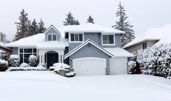A two-story blue house covered in snow with a white garage door, in a snowy neighborhood.