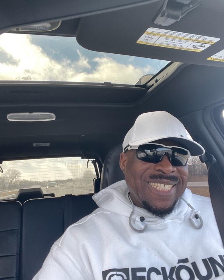 A smiling person wearing a white hoodie, sunglasses, and a white baseball cap inside a car with a sunroof.