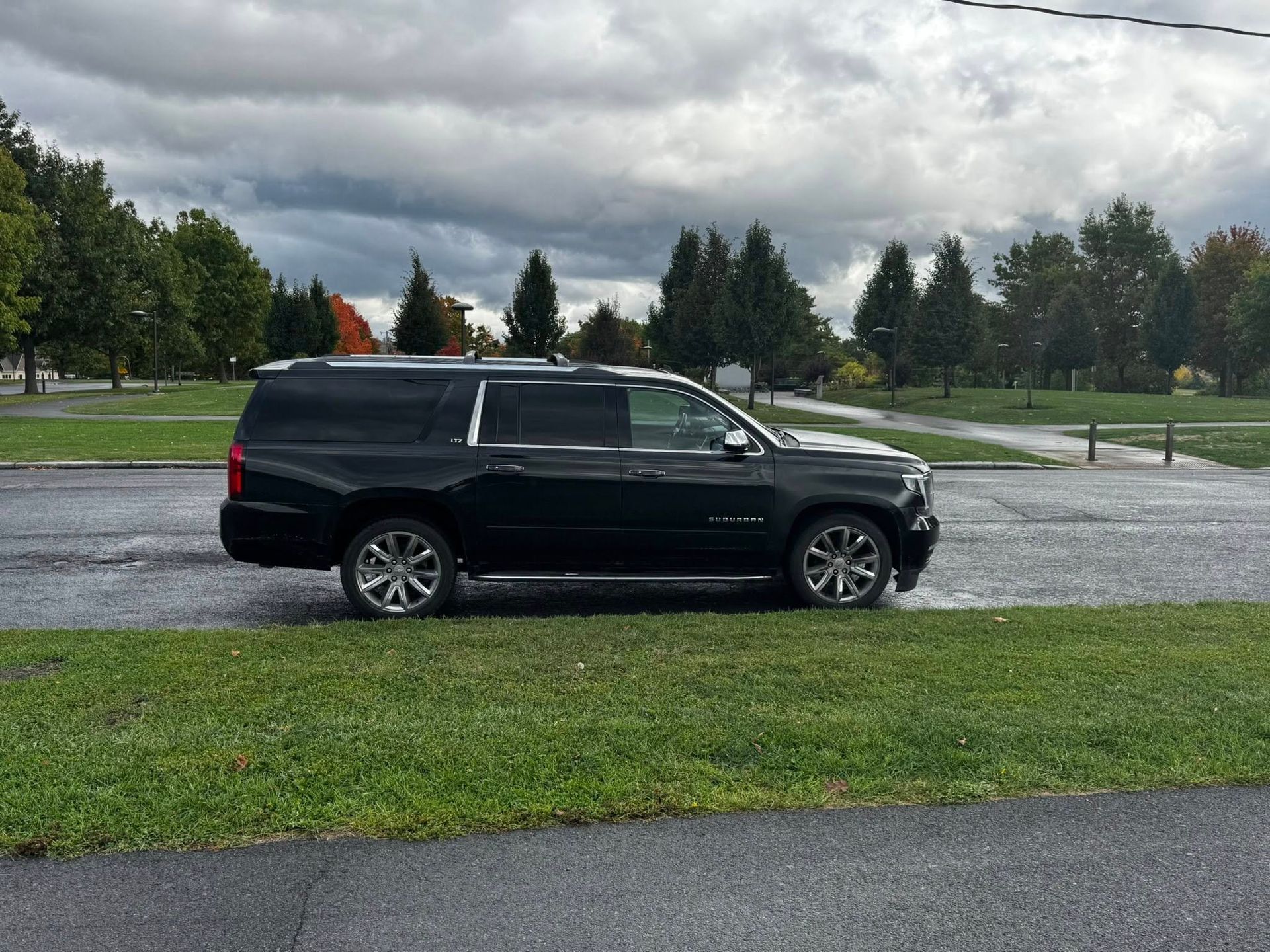 A black Chevrolet Suburban parked on asphalt next to a grassy lawn under a cloudy, overcast sky.