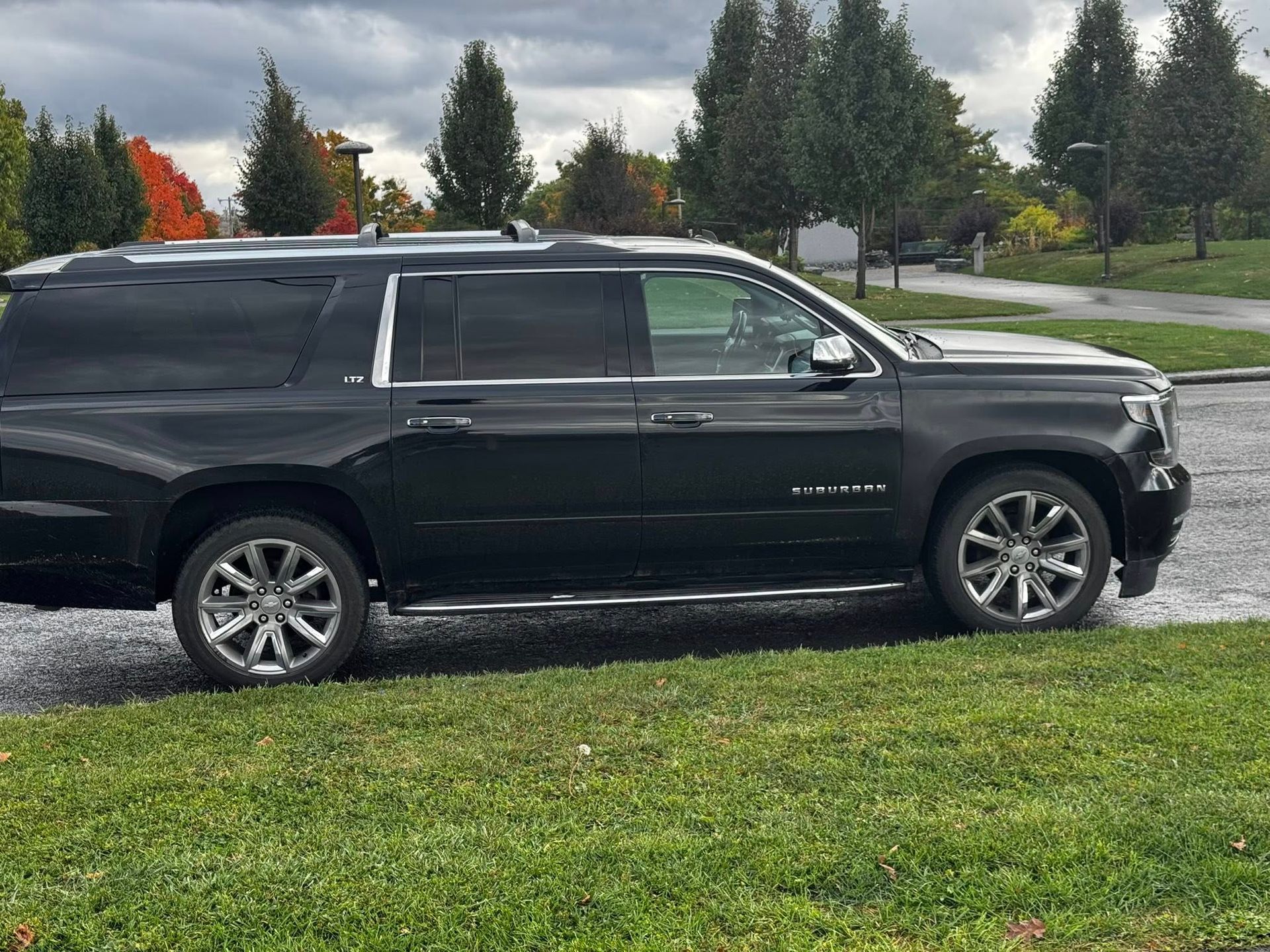 A black GMC Yukon SUV parked on a grass lawn next to an asphalt road, surrounded by trees under a cloudy sky.