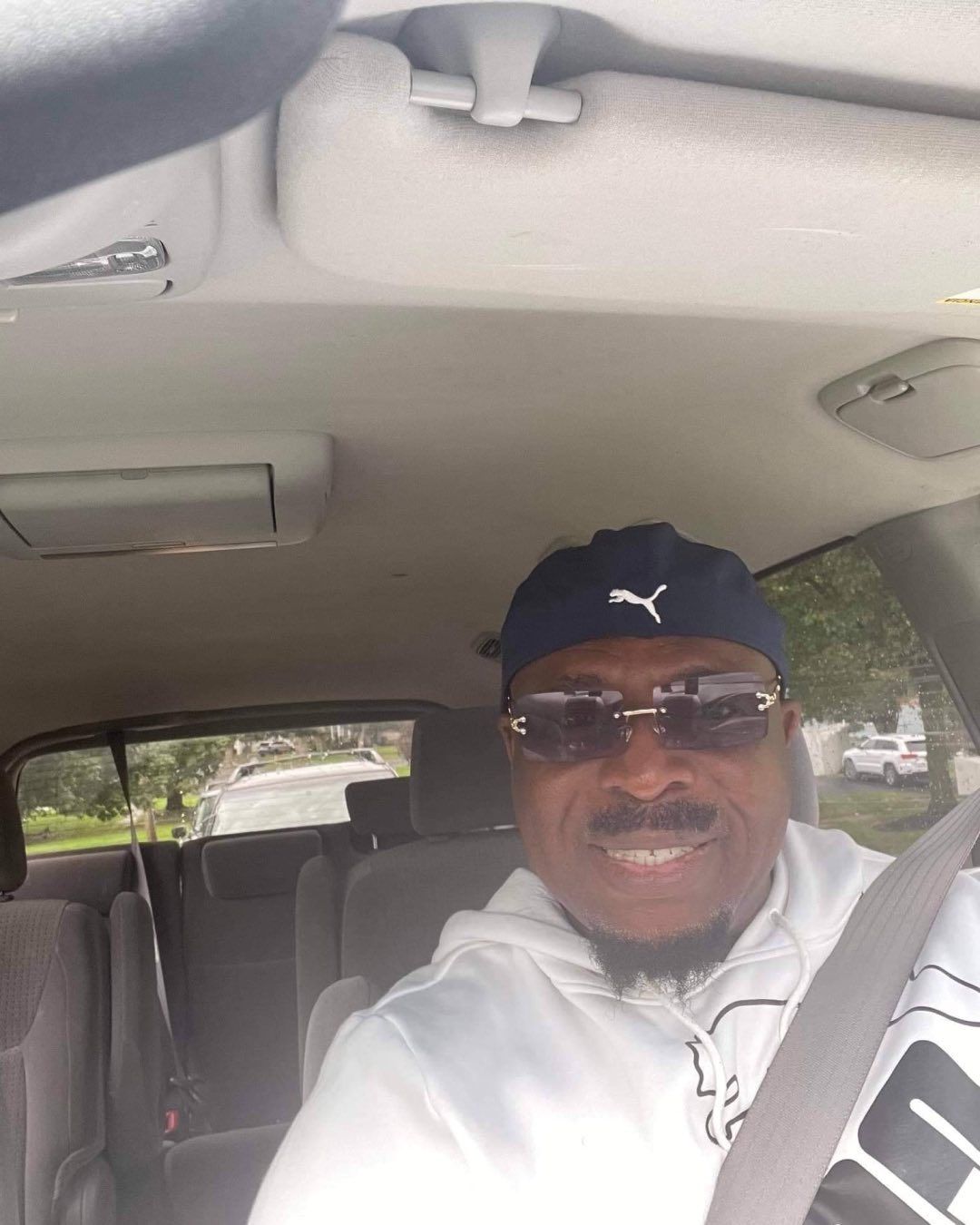 A man wearing a blue Puma cap, sunglasses, and a white hooded sweatshirt, smiling while sitting in a car.