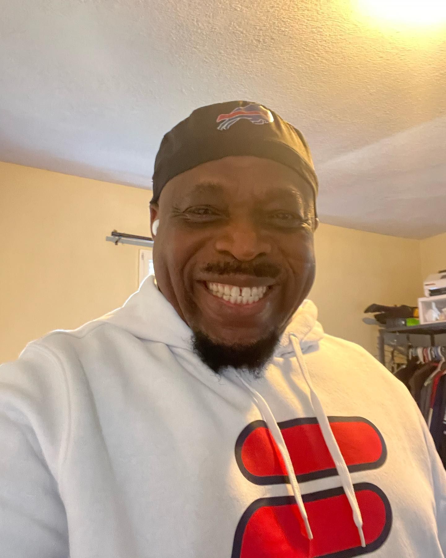 A smiling individual wears a dark baseball cap and a white hoodie with two large red horizontal bars on the chest.