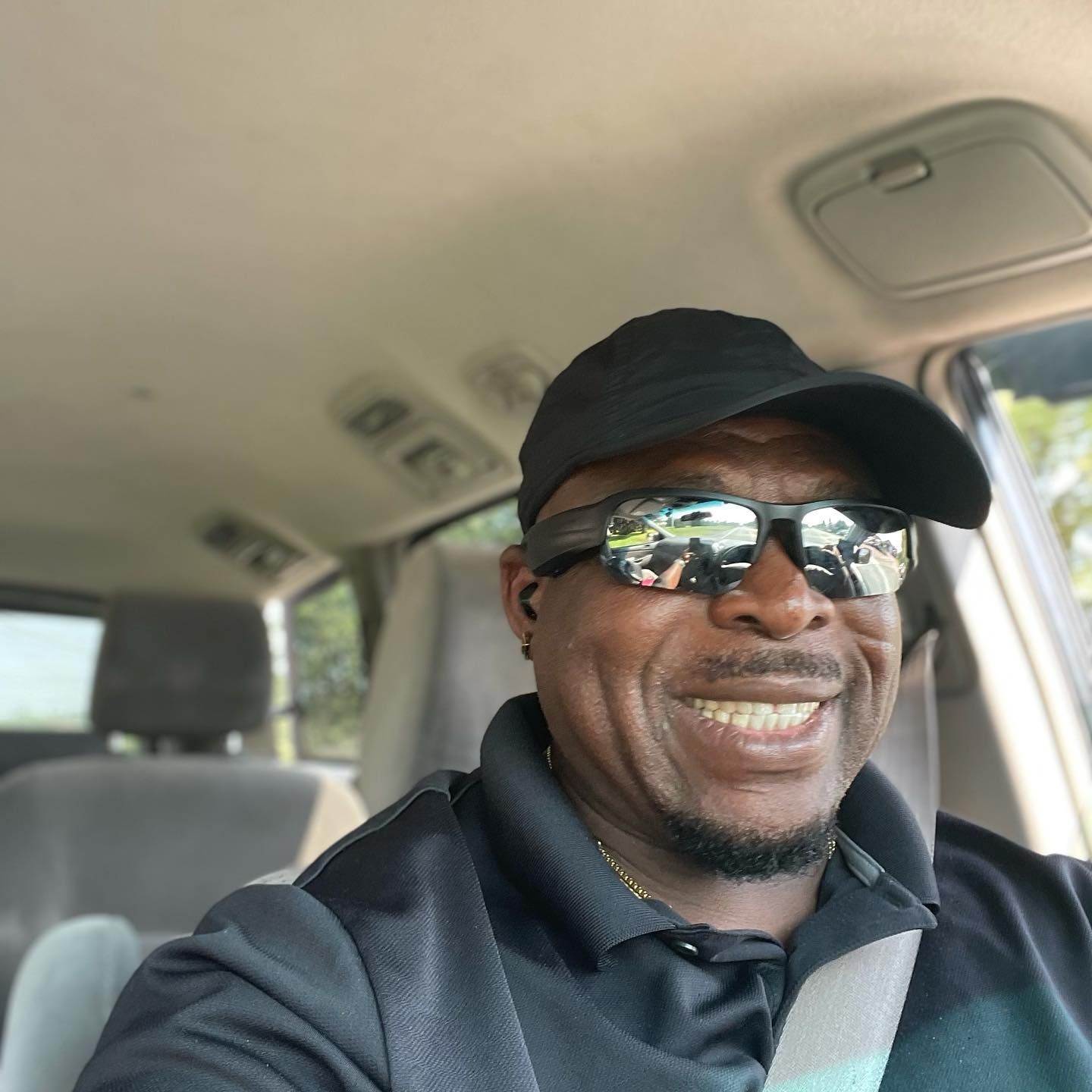 A smiling person wears a black baseball cap and dark sunglasses while sitting in the driver's seat of a car.