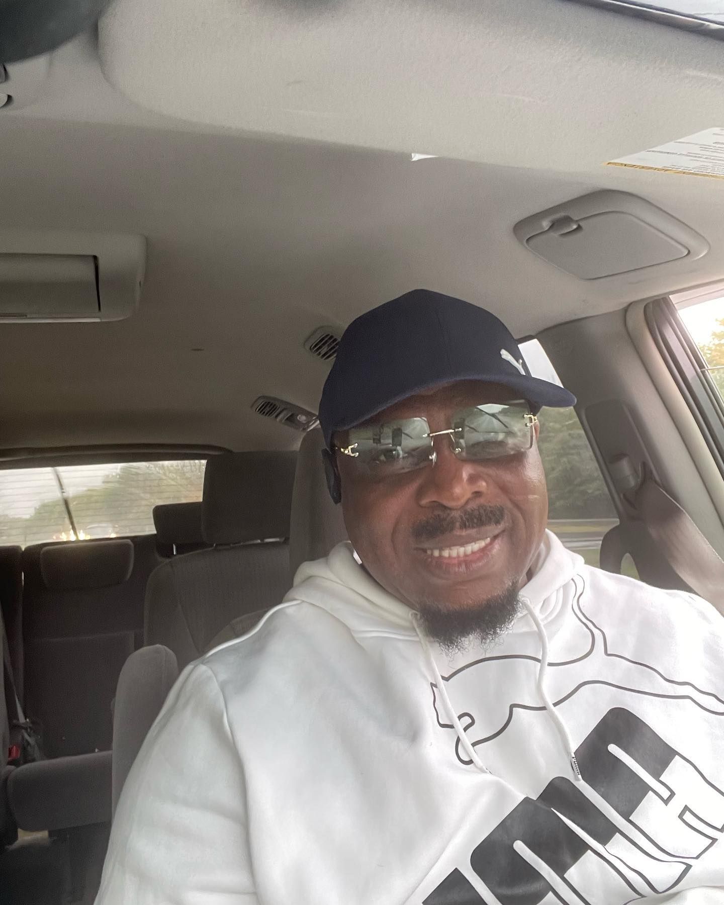 A man wearing a white Puma hoodie and a dark baseball cap smiles while sitting in the driver's seat of a car.