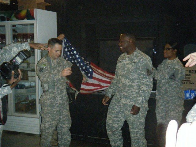 Three soldiers in camouflage uniforms stand before an American flag; one signs a document while another smiles.