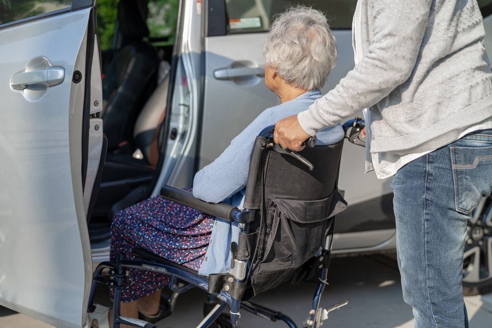 A person assisting an individual in a wheelchair to enter a silver car.