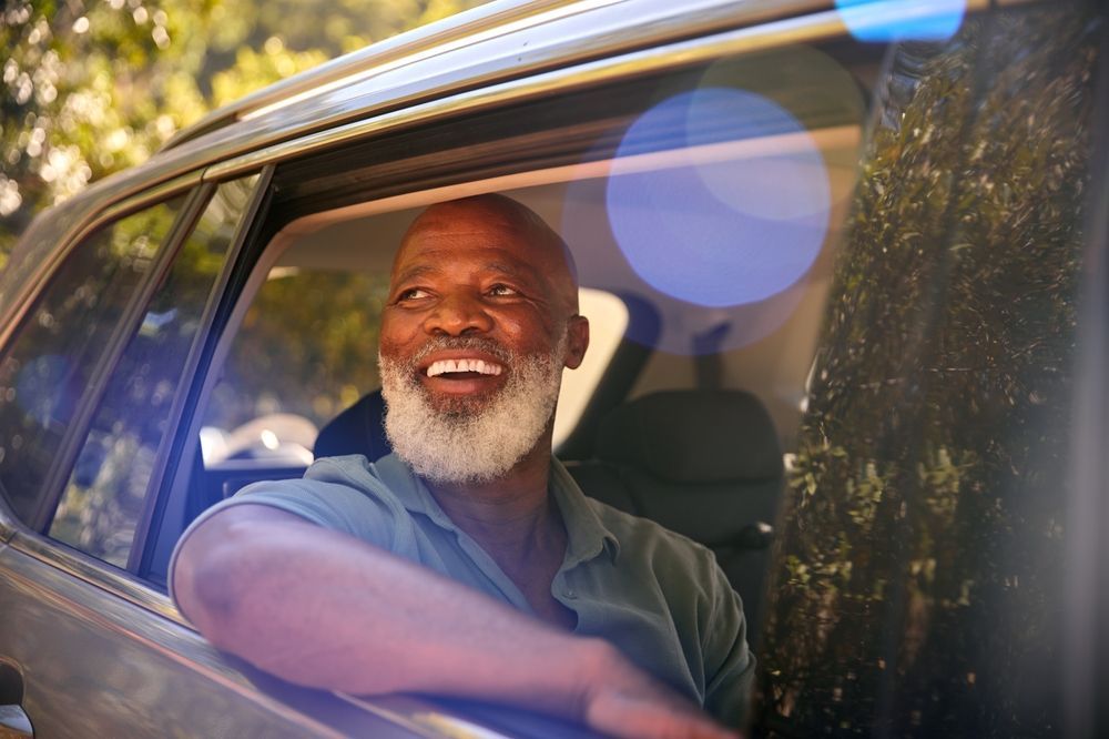 A smiling person with a grey beard looks out of a car window, leaning their arm on the frame on a sunny day.