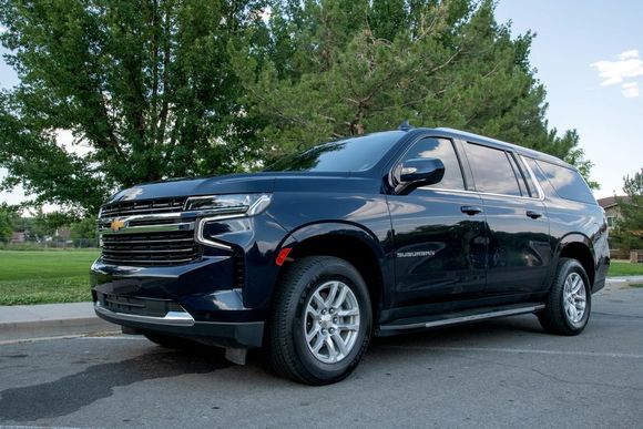 A dark blue Chevrolet Suburban parked on an asphalt road with green trees and a grassy field in the background.